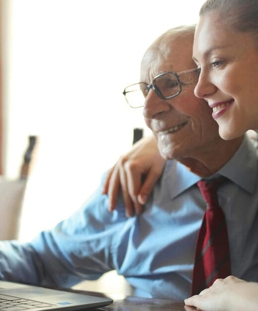 In the image, there's a heartfelt moment shared between a young woman and an elderly man. They seem to be family, perhaps granddaughter and grandfather. The young woman is leaning affectionately on the older man's shoulder, both of them smiling warmly while looking at a laptop screen. The man is wearing glasses, a light blue shirt, and a red tie, suggesting he may still be involved in professional activities or just prefers to dress smartly. The woman is dressed casually in a gray top, her hair tied back in a ponytail, giving her a relaxed and approachable look. The interaction looks positive, suggesting they are enjoying whatever is displayed on the screen, possibly sharing a digital experience or learning moment together.