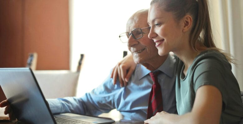In the image, there's a heartfelt moment shared between a young woman and an elderly man. They seem to be family, perhaps granddaughter and grandfather. The young woman is leaning affectionately on the older man's shoulder, both of them smiling warmly while looking at a laptop screen. The man is wearing glasses, a light blue shirt, and a red tie, suggesting he may still be involved in professional activities or just prefers to dress smartly. The woman is dressed casually in a gray top, her hair tied back in a ponytail, giving her a relaxed and approachable look. The interaction looks positive, suggesting they are enjoying whatever is displayed on the screen, possibly sharing a digital experience or learning moment together.